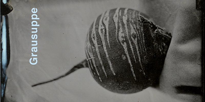 Ein dunkles, körniges Bild zeigt eine gestreifte, runde Knolle mit einem Stiel. Am linken Rand steht vertikal in weißer Schrift "Grausuppe".