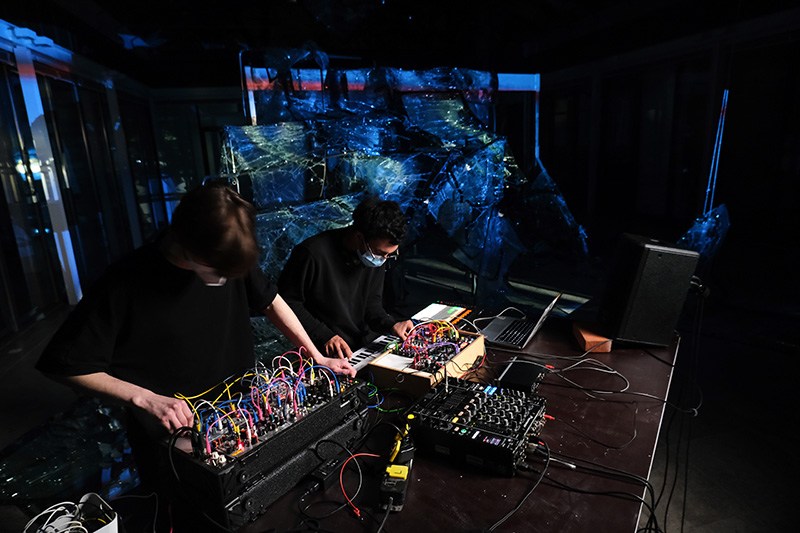 Zwei Personen arbeiten an elektronischen Musikgeräten und Laptops in einem dunklen Raum mit bläulich-reflektierenden Objekten im Hintergrund.