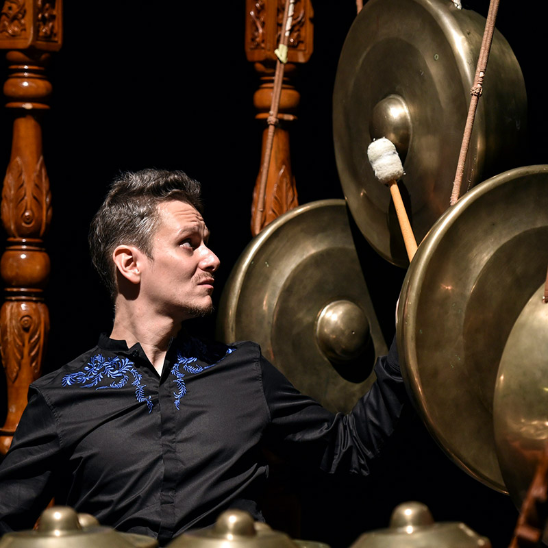 Ein Mann in schwarzem Hemd spielt mit einem Schlägel auf einem hängenden Gong aus einem traditionellen Gamelan-Ensemble.