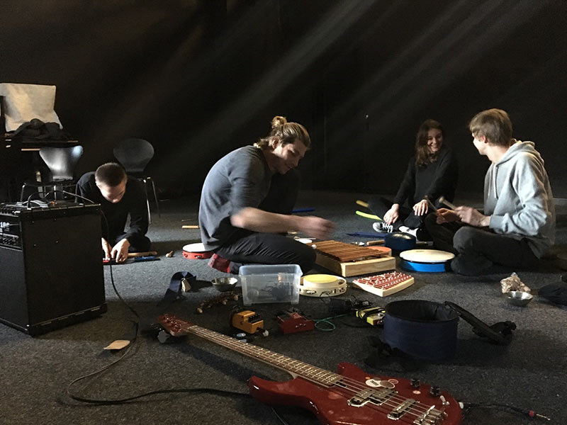 Vier Personen sitzen im Kreis auf dem Boden in einem abgedunkelten Raum und spielen verschiedene Musikinstrumente, darunter ein Xylophon, Trommeln und eine E-Gitarre. Ein Verstärker steht links.