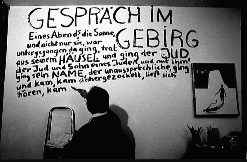 Ein Schwarz-Weiß-Foto zeigt eine Person, die auf eine Wand mit handgeschriebenem Text deutet, der mit "GESPRÄCH IM GEBIRG" beginnt und eine Passage über einen Juden enthält. An der