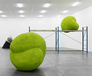 In einem weißen Ausstellungsraum stehen mehrere skulpturale Objekte, darunter zwei große, hellgrüne, organisch geformte Objekte, eines davon auf einem blauen Regalgestell.