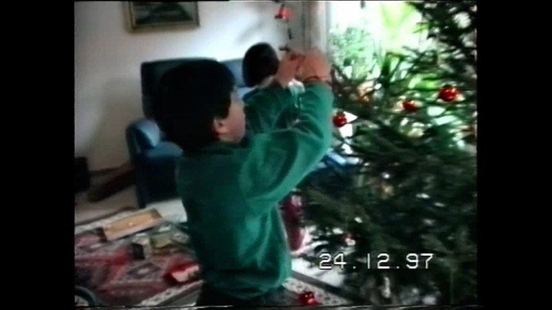 Ein unscharfes, körniges Video vom 24.12.97 zeigt zwei Kinder, die einen Weihnachtsbaum mit roten Kugeln und anderen Ornamenten schmücken.