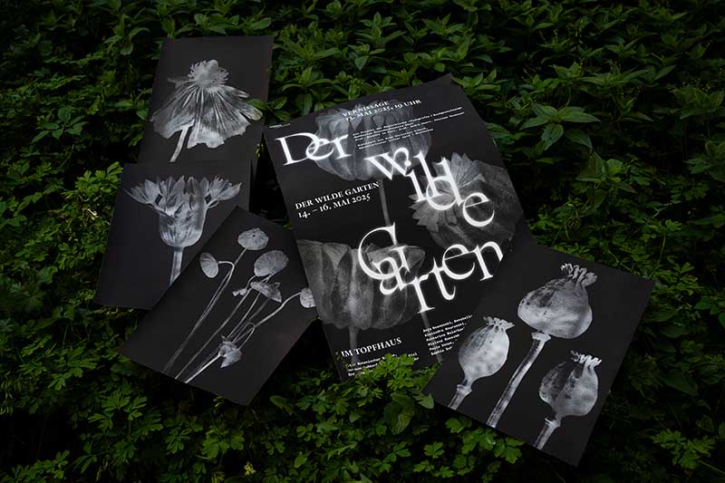 Mehrere schwarze Plakate mit weißen, teils verspielten Schriften und monochromen Pflanzenabbildungen liegen auf dichtem, grünem Laub. Im Zentrum steht ein Poster mit dem Titel „Der wilde Garten“, das eine Veranstaltung vom 14. bis 16. Mai 2025 im Topfhaus ankündigt. Die Gestaltung ist kontrastreich, künstlerisch und wirkt wie eine Mischung aus botanischer Illustration und moderner Typografie.