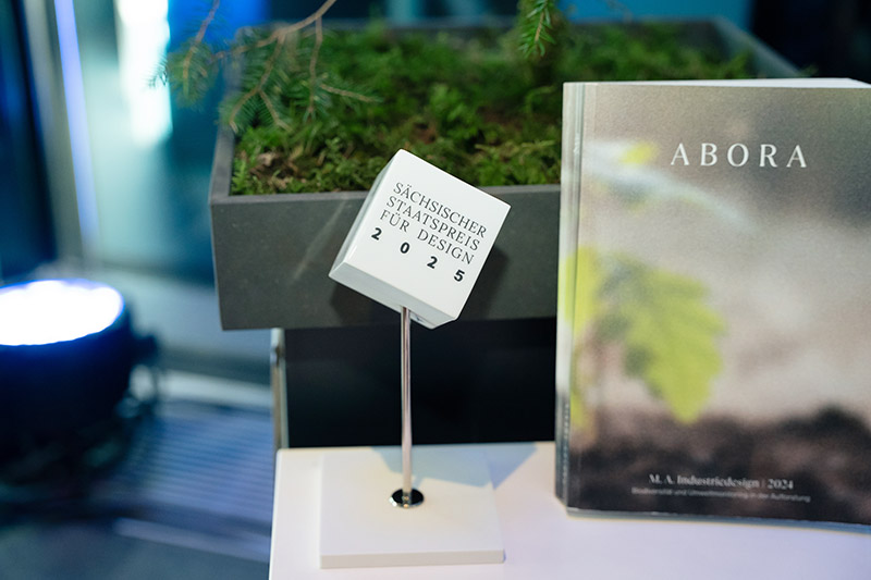 Auf einem weißen Tisch steht eine Auszeichnung in Form eines weißen Würfels neben einem Buch über Bäume